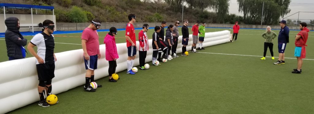 Athletes stand against the inflatable barricade, each with a soccer ball at their feet, while listening to instruction from coach Tim Taylor