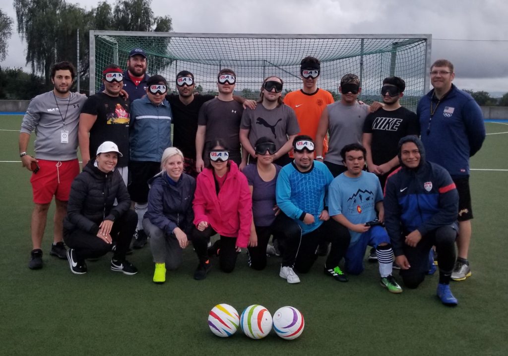 Athletes, coaches and staff pose for a group photo in front of a soccer goal. Three soccer balls are positioned on the ground in front of the group