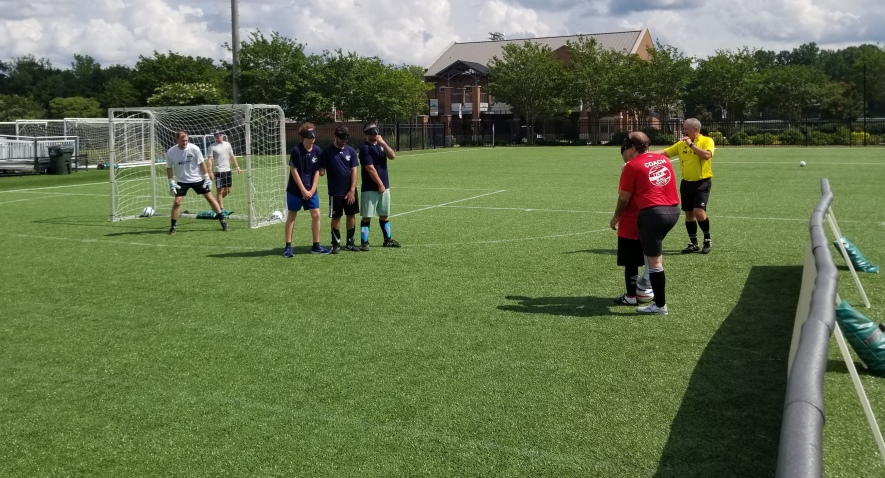 Cody Kirchner stands behind teammate Alvaro Mora with his foot on top of the soccer ball. Defenders on the other team form a wall in front of their goalkeeper.