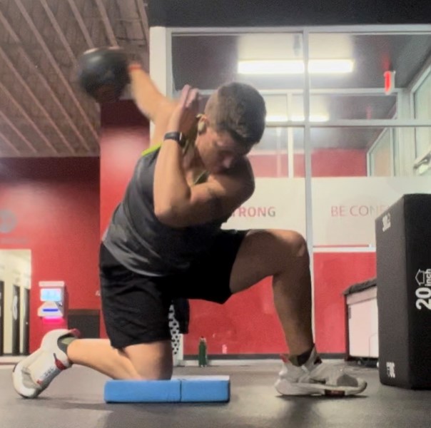Kneeling on her right knee atop a blue pad, Asya swings her right arm back while holding a small weighted ball.