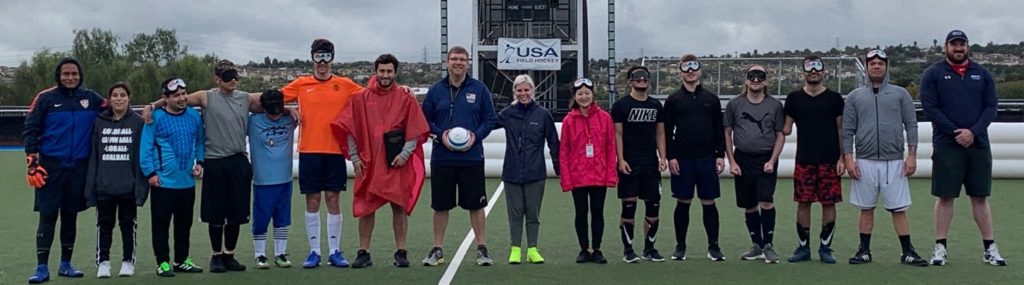 Athletes, coaches and staff stand shoulder-to-shoulder, some with their arms around each other, on the soccer pitch during the talent ID camp
