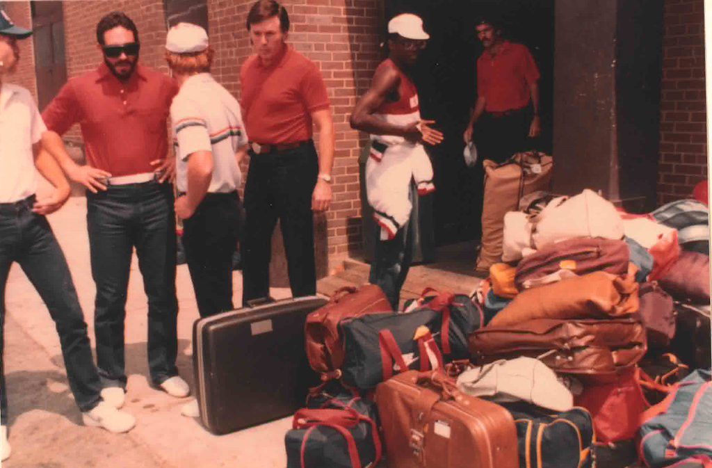 Athletes and officials look over a pile of luggage on an outside sidewalk.