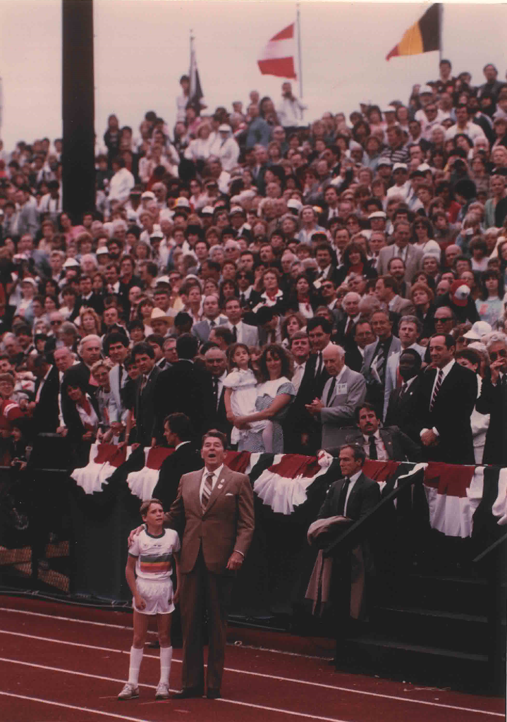 U.S. President Ronald Reagan stands on the track with his arm around a young boy with a bleacher full of spectators in the background