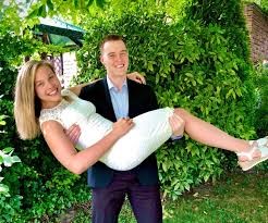 Amanda's husband Michael carries Amanda in his arms on their wedding day in 2020. Amanda is wearing a white dress and Michael is in a dress shirt and blazer. Green trees and bushes are in the background.