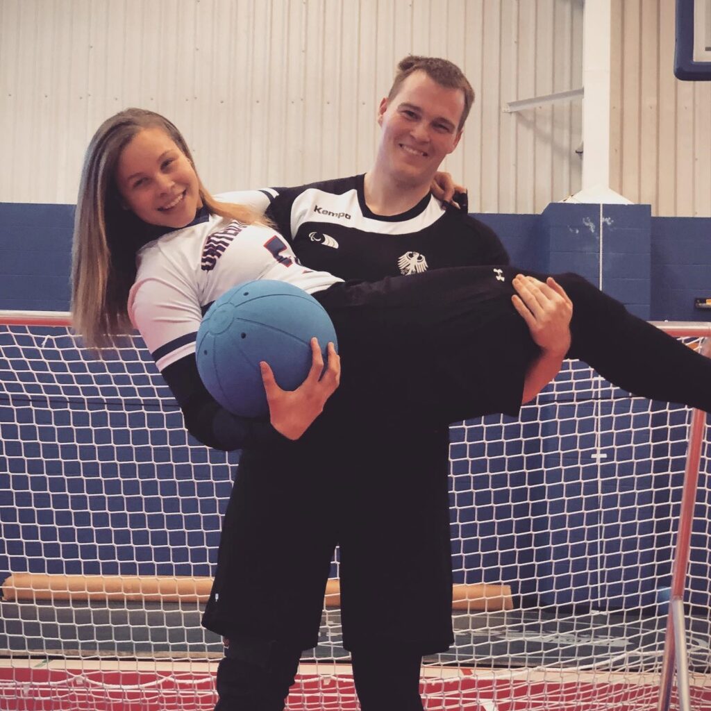 Amanda is held in the arms of her husband Michael as they pose on the goalball court in front of a goal. Amanda holds a goalball in her right hand.