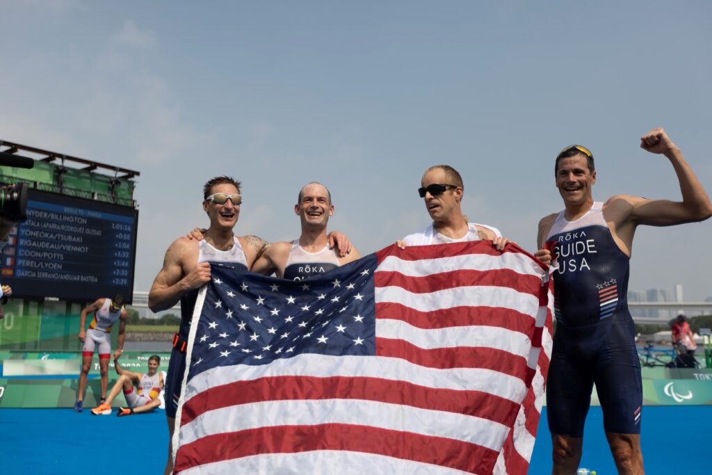 The Tokyo 2020 U.S. Paralympic Team holds an American flag in front of them following the men's VI triathlon race in Tokyo in 2021. Pictured are Brad Snyder and his guide Greg Billington, and Kyle Coon and his guide Andy Potts.