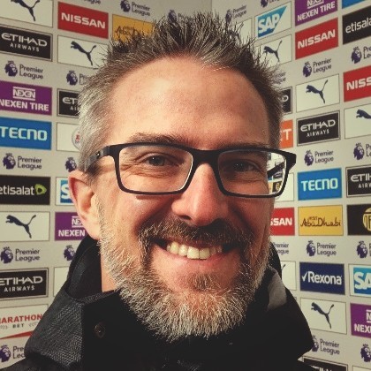 A headshot of Brian Eaton smiling and standing in front of a wall of sponsor logos. Brian sports a beard and is wearing glasses.