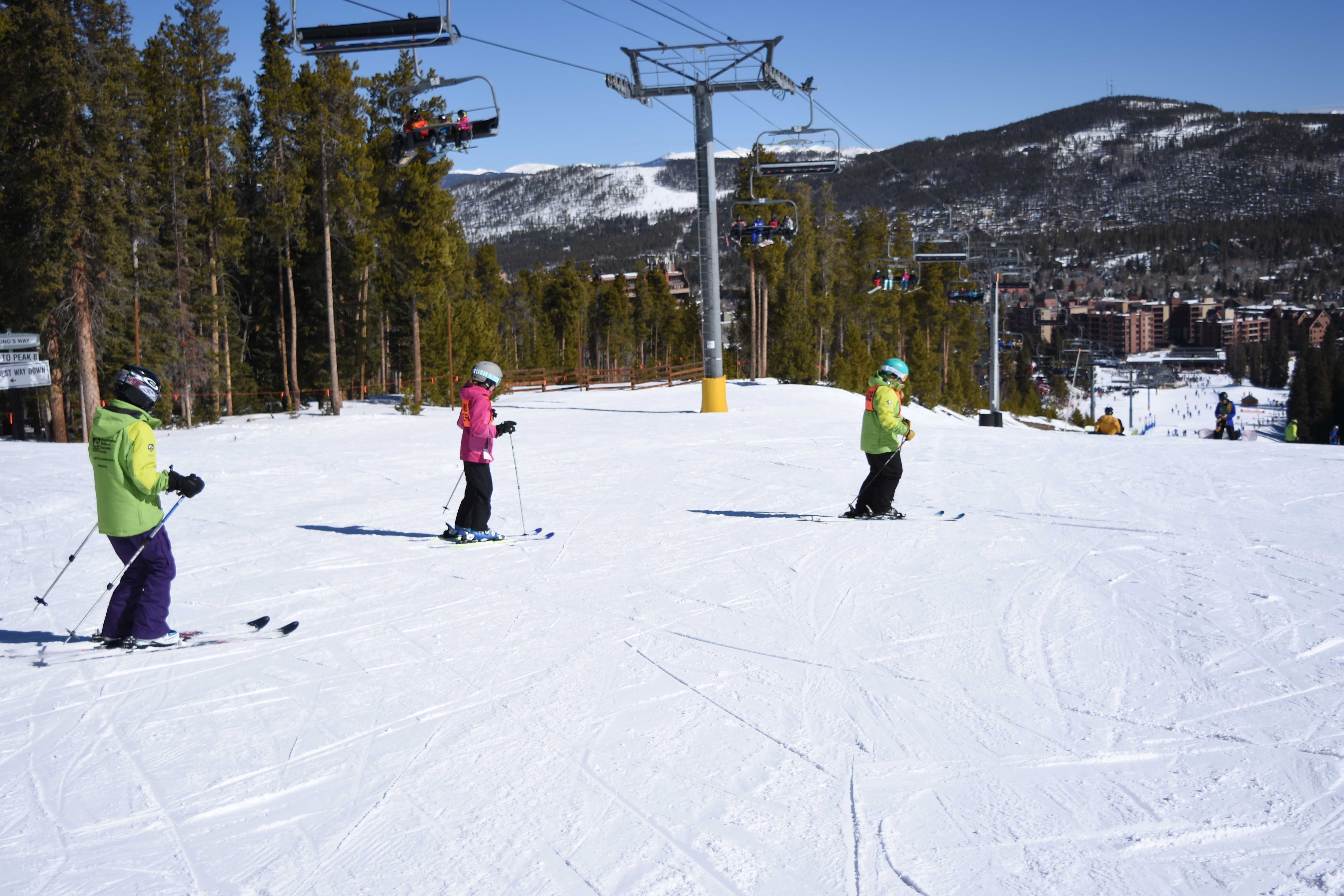 In a bright pink jacket, Caroline follows a guide wearing a green jacket toward the bottom of the hill in front of them. Another guide in a green jacket follows behind Caroline. There is a ski lift and trees to their left and open space to their right.
