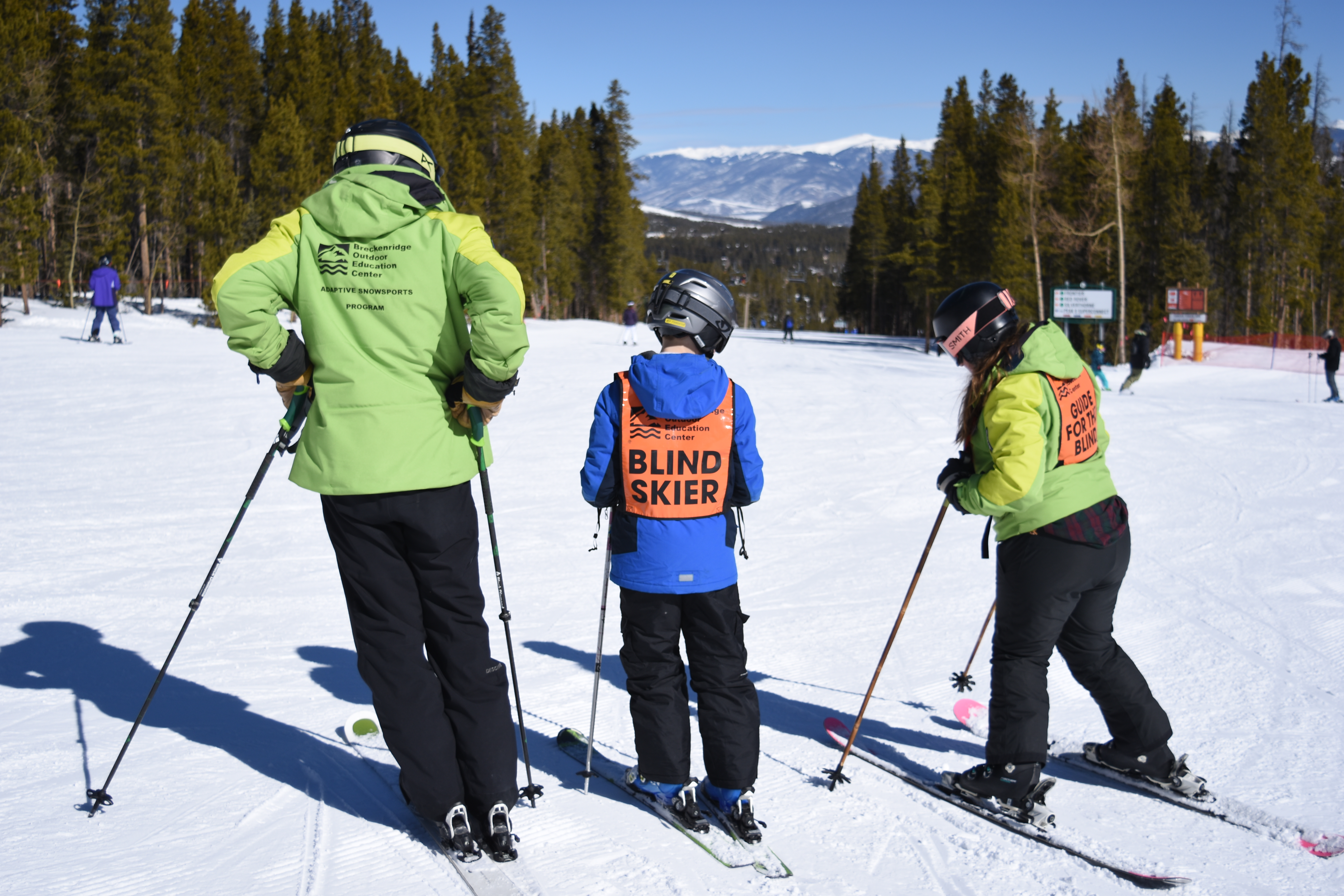 Cole Carper wears an orange blind skier vest over his bright blue jacket and stands between his two ski guides. They stand at the top of a run they are about to ski down. Trees and other skiers around them and snowcapped mountains in the distance.