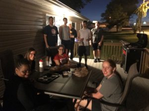 Resident goalball players sitting on deck outside a house, some are sitting around a table and some are standing. They are enjoying a cookout.