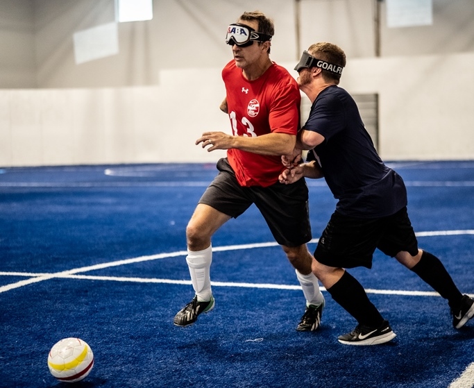 Kevin Brown and Noah Beckman battle for control of the soccer ball. Brown is wearing a red jersey and Beckman is wearing blue. Both have eyeshades on.