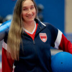 Eliana Mason poses with a goalball under her left arm and wearing a USA Goalball blue jacket