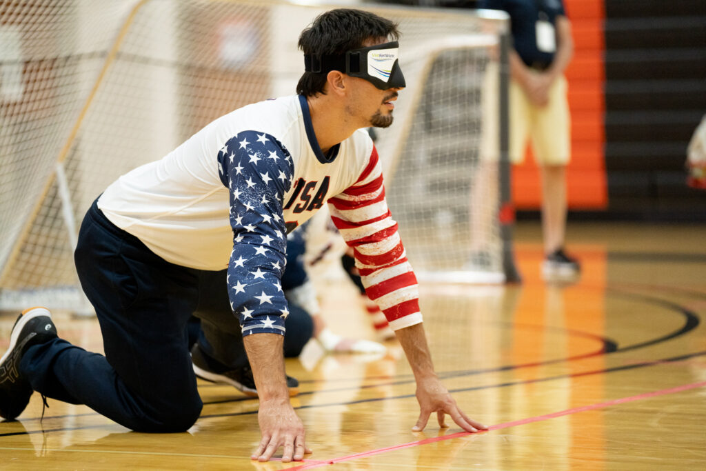 Tyler Merren leans on his right knee with his fingertips touching the floor as he anticipates an opponent's throw during the 2019 IBSA International Goalball Qualifier in Fort Wayne, Indiana. (photo by Julie Larame)