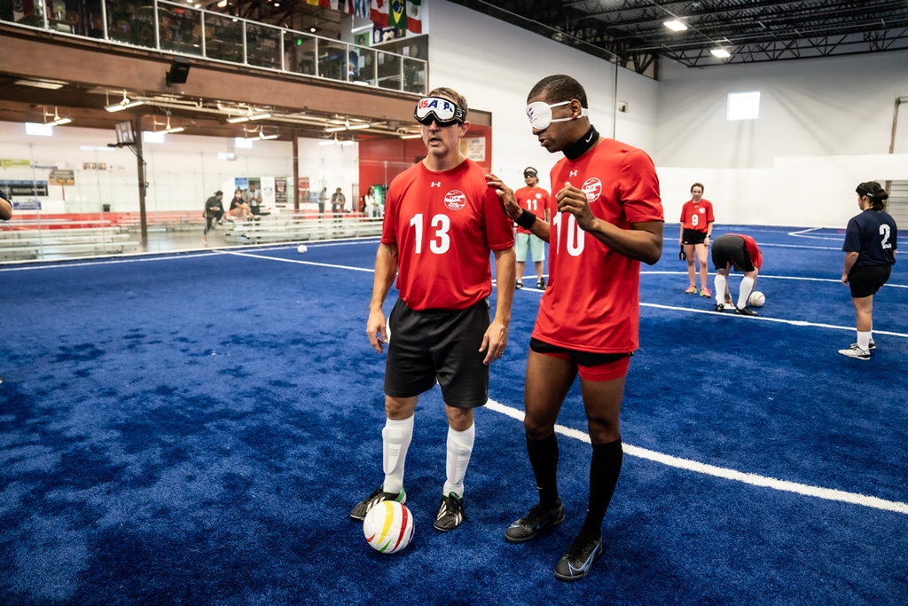 Wearing red USA Blind Soccer jerseys and eyeshades, Kevin Brown (l) and Antoine Craig practice putting the ball in play. Craig has his right hand on Brown's left shoulder as he stands next to him. A soccer ball is at Brown's feet.