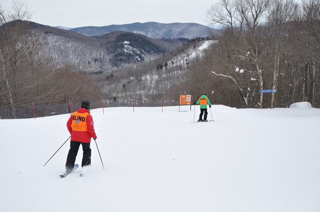 A skier wearing an orange BLIND vest follows their guide down the slopes on Pico Mountain.