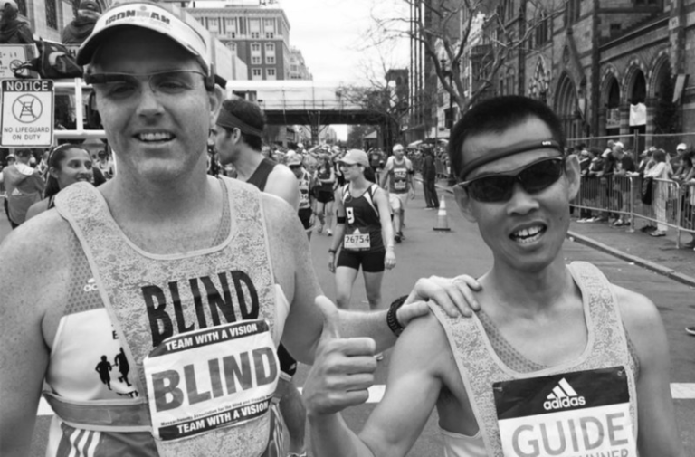 Black and white photo of Erich Manser and his guide. Erich is on the left and has his left hand on the shoulder of his guide who is standing beside Erich wearing sunglasses and giving the camera a thumbs up.