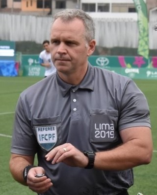 Skye Arthur-Banning is pictured officiating at the 2019 Parapan American Games in Lima, Peru. He is wearing a gray shirt with a white patch over the right breast reading "Referee IFCPF" and the "Lima 2019" insignia on the left breast.