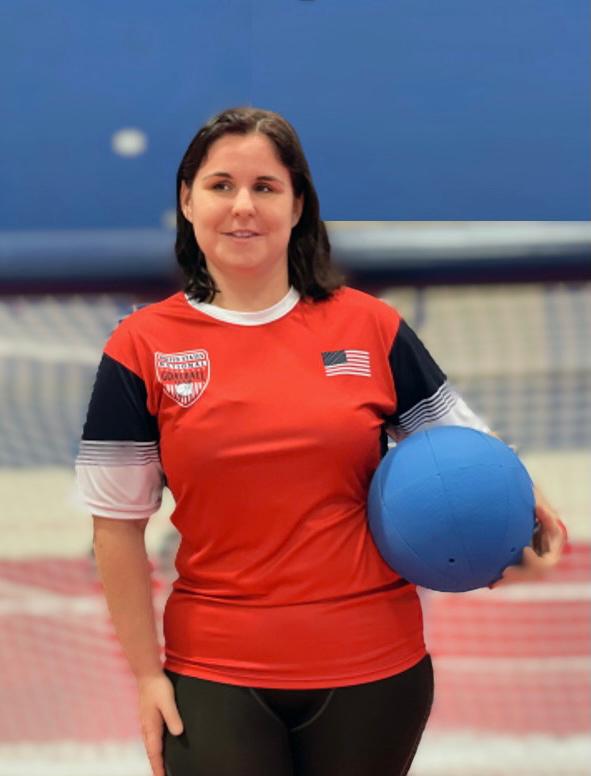 Libby Daugherty poses while holding a goalball in her left hand. She is wearing a red USA Goalball short sleeve shirt with a USA flag on the left chest.