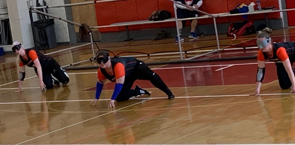 Libby plays center between two teammates as they anticipate a throw from their opponent during a tournament. They are wearing eyeshades, black pants and and black jerseys with orange sleeves.