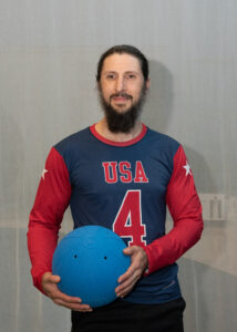 A photo of John Kusku holding a goalball and wearing his USA jersey with the number 4 on the front.