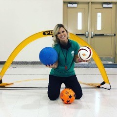 Katie Smith kneels in front of a goal holding a goalball in her right hand and a soccer ball in her left hand. Another soccer ball is on the ground in front of her.