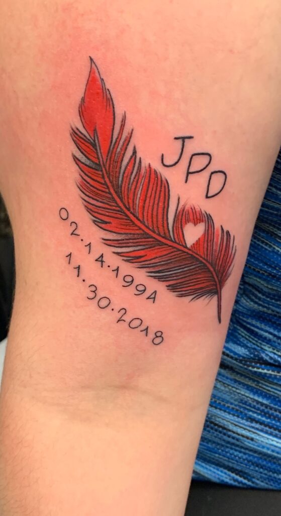 A red cardinal feather is shown tattooed on Libby Daugherty's arm. Above the feather in black ink are the initials JPD. Below the feather in black ink are the dates 02.14.1994 and 11.30.2018.