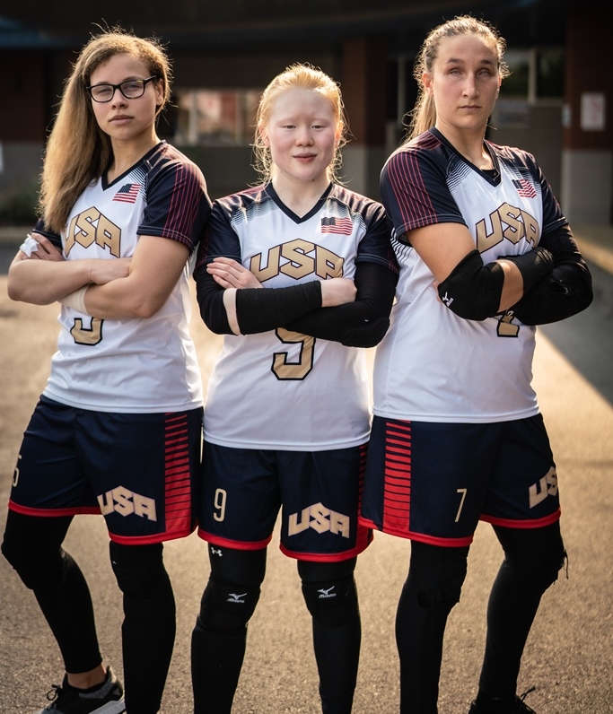 Marybai Huking stands between teammates Amanda Dennis and Eliana Mason. All three have their arms folded with serious expressions on their faces. They are wearing white USA jerseys and blue shorts with gold lettering and numbers.