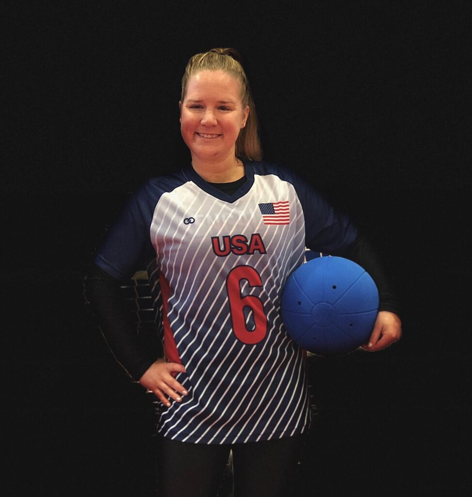 Mindy Cook poses for a photo with a goalball under her left arm. She is wearing a Team USA jersey with #6 on the front.