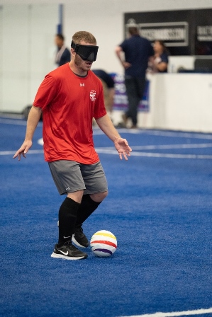 Wearing a red t-shirt and black eyeshades, Noah Beckman dribbles the ball down the soccer pitch which has blue turf.