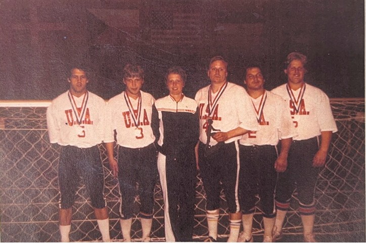 The 1980 USA Goalball men's team poses with their silver medals alongside assistant coach Eugenie Kriebel in front of a goal.