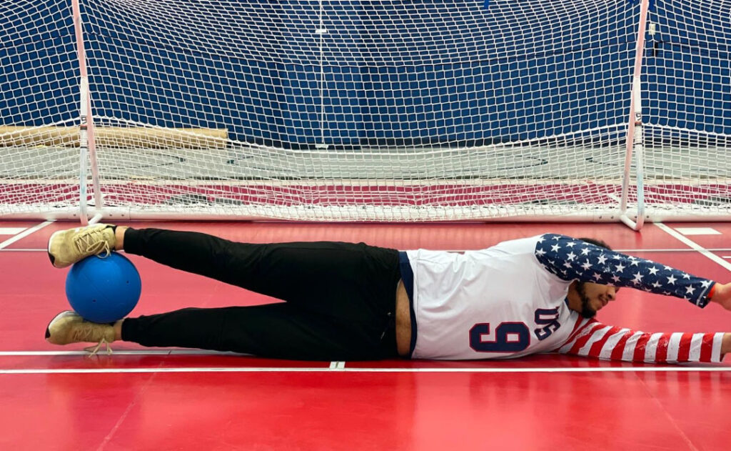Sean Walker stretches out horizontally to stop the goalball with his feet in front of a goal. He is wearing a USA #9 jersey and black pants.