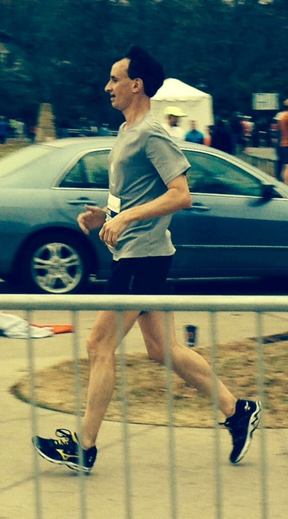 William runs past barricades on a sidewalk during a race. He is wearing a gray t-shirt, dark shorts and dark running shoes.