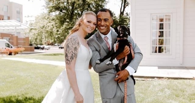 Zach and his wife Bailey pose on their wedding day. Zach is wearing a gray suit with pink tie and is holding a black and white puppy, while Bailey is wearing a white wedding dress.