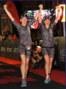 Randi Strunk and her guide finishing the Ironman Texas in 2018. Both have their arms raised in the air, grinning.